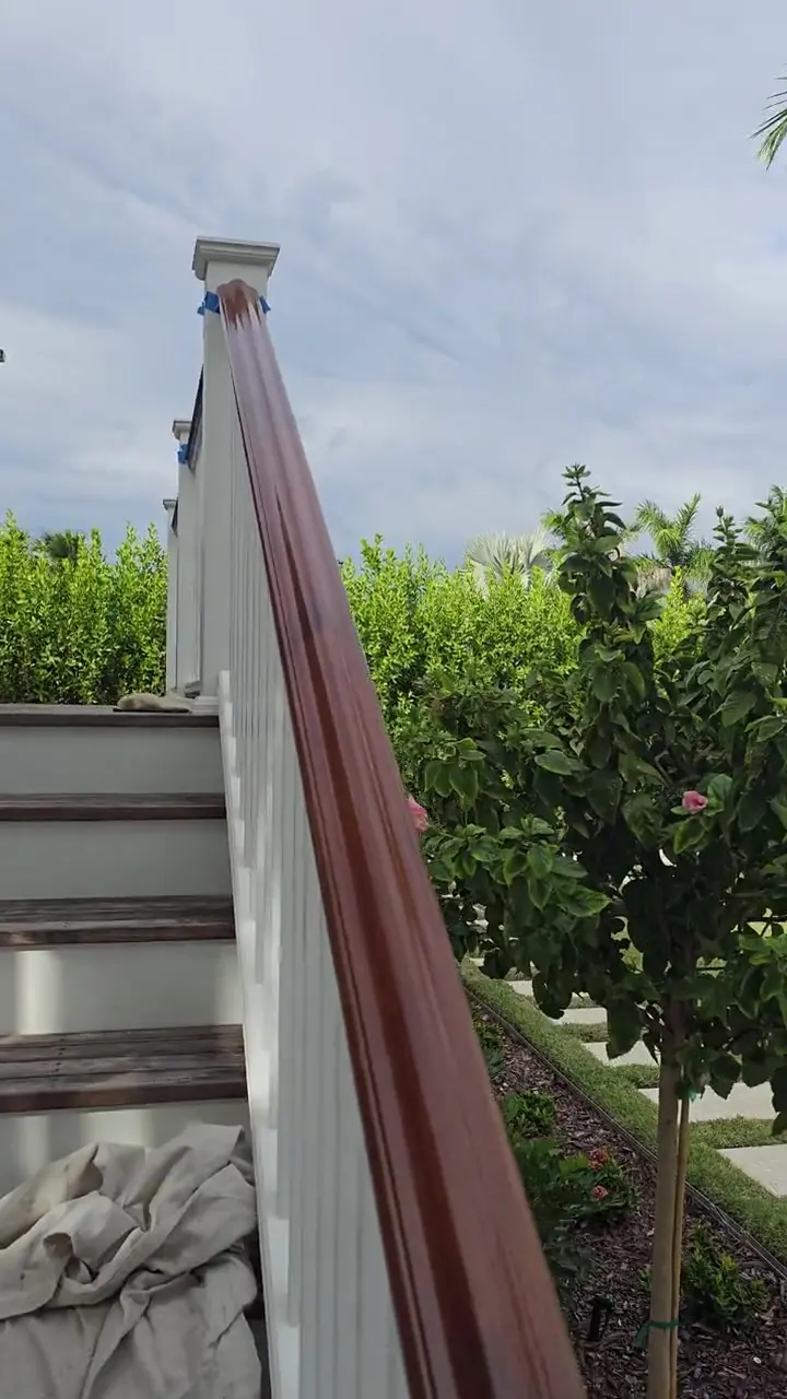 An exterior stair handrail finished in a warm mahogany stain over a white-painted balustrade — outdoor wood carrying a stain finish through Florida sun and rain.