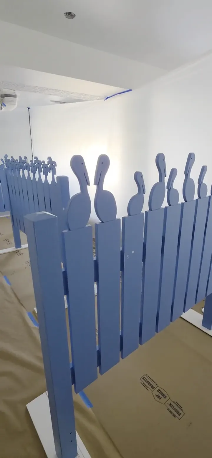 Cut-out pelican picket panels standing on drop cloths in the shop, carrying their first coat before the final white goes on.